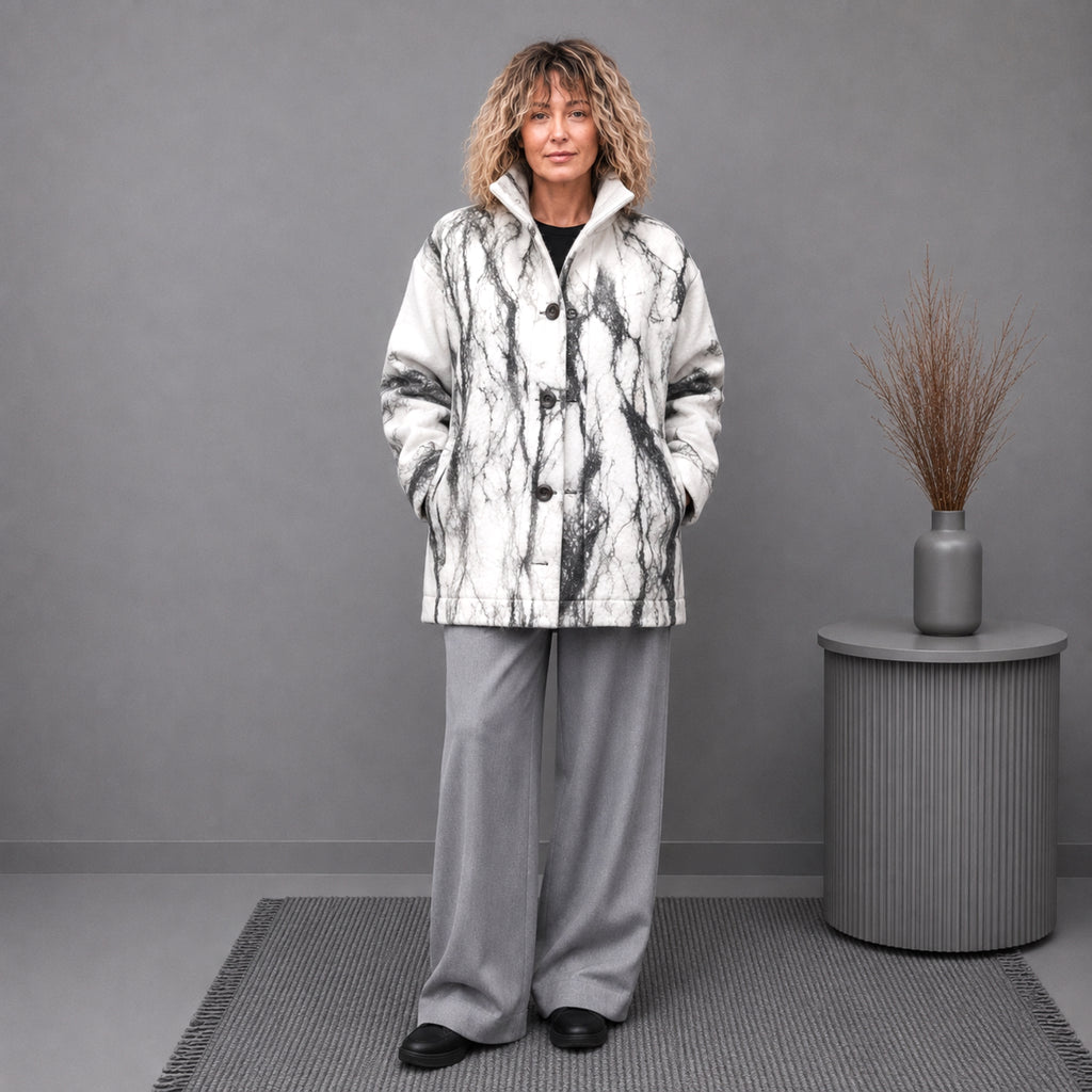 A woman stands in a studio setting wearing a white and black marble-patterned coat over a black top, paired with light gray wide-leg trousers and black shoes. A gray side table with a vase of dried grass is to her right against a plain gray wall.
