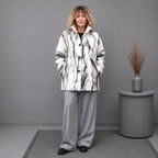 A woman stands in a studio setting wearing a white and black marble-patterned coat over a black top, paired with light gray wide-leg trousers and black shoes. A gray side table with a vase of dried grass is to her right against a plain gray wall.